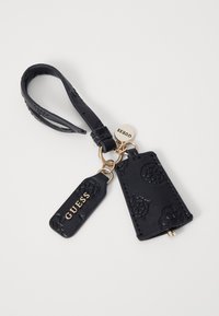 Portachiavi in pelle nera texturizzata con due etichette, una delle quali è impressa con "GUESS" in oro, attaccata da un anello e una cinghia in metallo dorato.