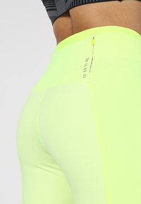 Leggings ajustados de color amarillo neón con una tela texturizada, que cuentan con un bolsillo con cremallera en la parte posterior y números impresos a lo largo de la costura.