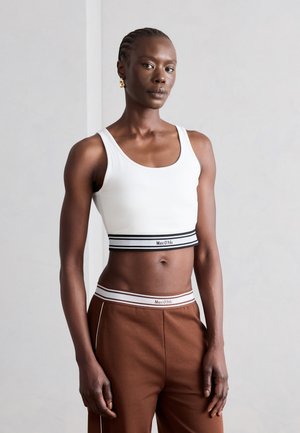 Γυναίκα με πλεξούδες φοράει λευκό crop top και καφέ παντελόνια, και τα δύο με ελαστικές μέσες με λογότυπο, όρθια μπροστά από απλό φόντο.