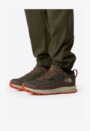 Baskets vertes et marron avec une tige en mesh, des accents orange et une semelle coussinée. Comprend un logo et des détails texturés.