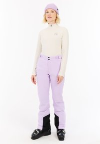 Paarse sneeuwbroek met zwarte gaiters, beige langslouw top met halve rits en logo, en een lichtpaarse gebreide beanie.