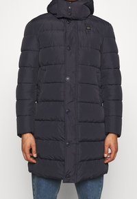 Marinblå pufferjacka med huva, med horisontell söm, framknappar och sidofickor. Slät, vattenavvisande tyg, längd till mitten av låret.