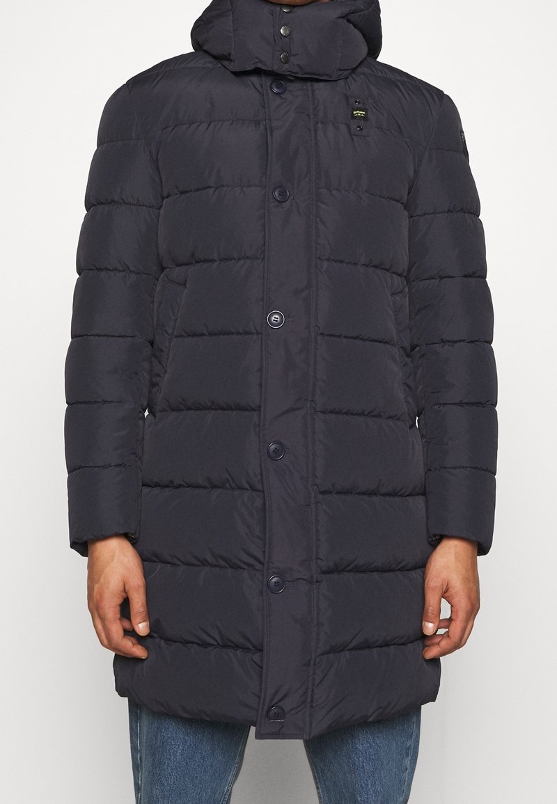 Marinblå pufferjacka med huva, med horisontell söm, framknappar och sidofickor. Slät, vattenavvisande tyg, längd till mitten av låret.