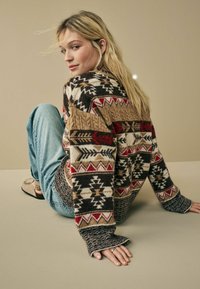 Pull en tricot avec un motif géométrique marron, crème, rouge et noir, doté de manches côtelées et d'une coupe décontractée, associé à un jean bleu.