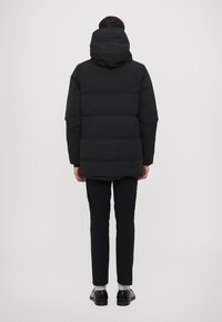 Geacă puffer neagră cu glugă, având un design matlasat. Purtați-o peste pantaloni bleumarin și încălțăminte neagră. Estetică simplă, minimalistă.