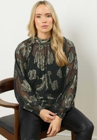 Blusa nera trasparente con motivo floreale metallico, collo alto e maniche ampie. Abbinata a pantaloni in pelle nera aderenti.
