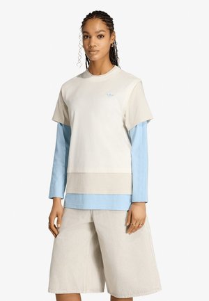 Donna con capelli intrecciati che indossa una camicia a strati beige e azzurro a maniche lunghe e pantaloni beige a gamba larga fino al ginocchio, su sfondo bianco.