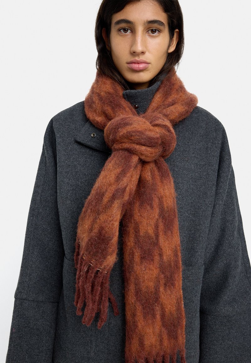 Wełniany szal w ciepłym, rudobrązowym kolorze o miękkiej fakturze, z frędzlami na końcach i węzłowym wzorze, noszony na ciemnoszarym płaszczu.