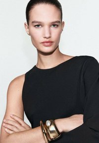 Top negro sin mangas con un escote amplio; el modelo lleva una pulsera de oro gruesa con un gran acento de perla, piel suave y cabello liso.