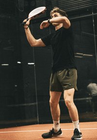 Jonge man in zwart shirt en groene shorts speelt paddle-tennis binnenshuis en maakt zich klaar om de bal te slaan met een paddle-racket.