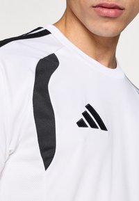 Λευκό αθλητικό πουκάμισο με μαύρες λεπτομέρειες στους ώμους. Διαθέτει αεριζόμενο ύφασμα με υφή και μαύρο λογότυπο με τρεις ρίγες.