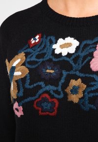 Pull noir tricoté avec broderie florale colorée en rouge, bleu, rose, jaune et blanc, agrémentée de fils bleus entrelacés.