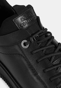 Baskets en cuir noir avec des accents en tissu texturé, lacets ronds et œillets en métal. Présente un logo en relief sur la languette et des détails brodés.