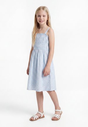 Νεαρό κορίτσι με μακριά ξανθά μαλλιά που φοράει ανοιχτομπλε αμάνικο φόρεμα με κόκκινες βούλες και άσπρα σανδάλια, στέκεται και χαμογελά.