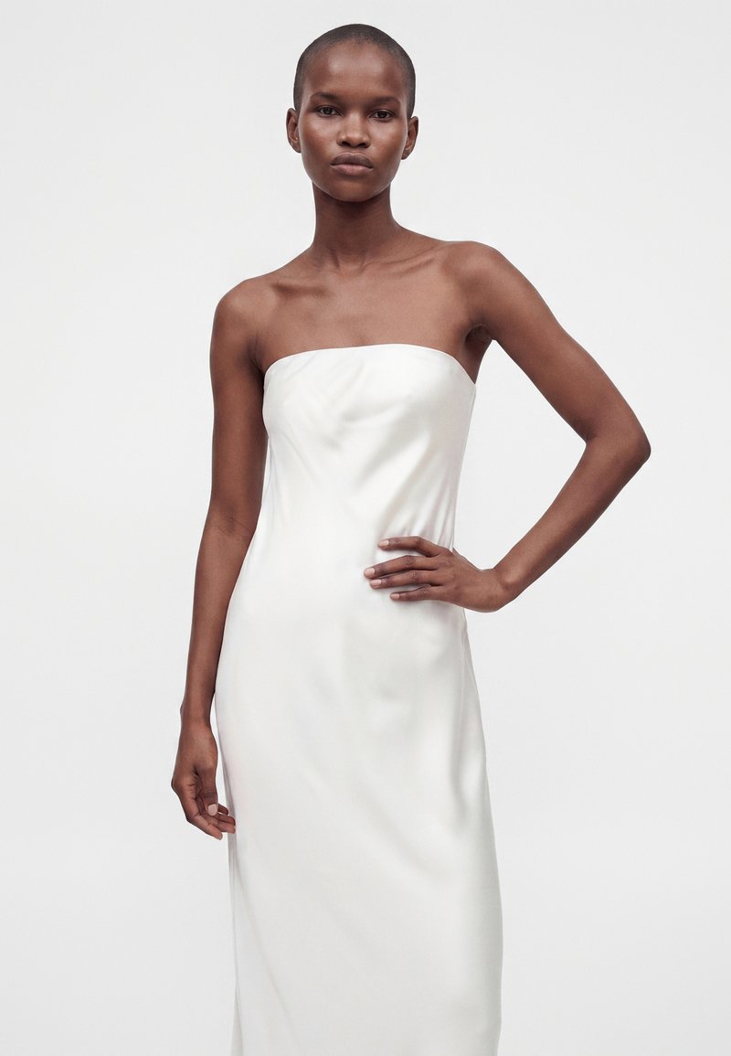 Femme aux cheveux courts portant une robe bustier en satin blanc, debout avec une main sur la hanche sur un fond uni.