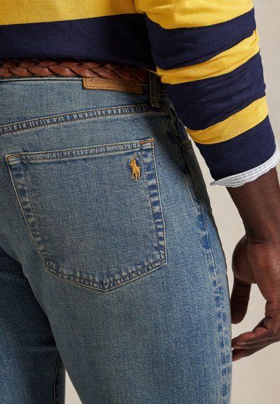 Mand iført lyseblå jeans med et brunt flettet bælte, gul og marineblåstribet skjorte, der viser baglommen med lille broderet logo.