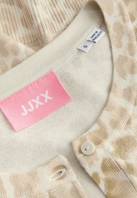 Gros plan sur l'encolure d'un cardigan beige à motif avec un bouton et une étiquette de marque rose JJXX sur un tissu tricoté doux.