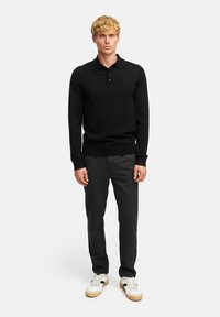 Polo shirt nero a maniche lunghe con colletto, abbinato a pantaloni grigio scuro e sneakers bianche con accenti neri e suola color gomma.