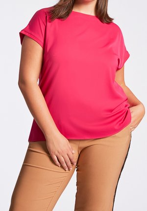 Vrouw draagt een felroze top met korte mouwen en beige broek, staat met één hand in de zak en de andere rustend op haar dij.