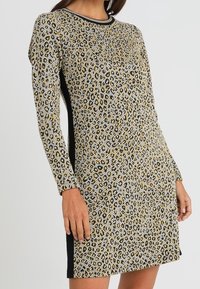 Robe à imprimé léopard avec des manches longues, motif doré et noir, encolure ronde et panneaux latéraux noirs. Fabriquée en tissu texturé et extensible.
