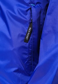 Tissu bleu avec une poche verticale zippée étiquetée "PACK POCKET" sur une veste ou un sac léger.
