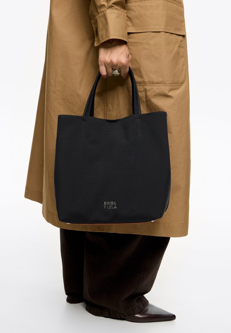 Zwarte tote bag met twee handvatten, gemaakt van textuurstof. Heeft een ingetogen logo op de voorkant en een eenvoudige, minimalistische vormgeving.