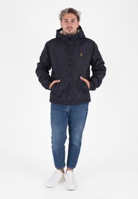 Giacca nera isolata con cappuccio, chiusura con zip e tasche frontali. Indossata con jeans azzurri chiari e sneaker bianche. Design e vestibilità casual.