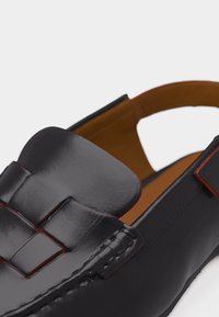 Μαύρος δερμάτινος ανδρικός μοκασίνι με πλεκτή λεπτομέρεια στο λουράκι και καφέ μαλακό εσωτερικό, απεικονίζεται σε γωνία σε λευκό φόντο.