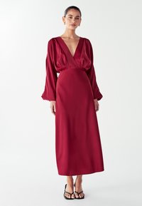 Burgundi aksominė maksi suknelė su giliu V formos iškirpimu, pūkuotomis rankovėmis ir prigludusia juosmeniu. Pasižymi lygiu tekstūru ir pilno ilgio dizainu.