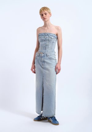 Ung person med kort blondt hår iført en stropløs denimkorsetter, lang denim nederdel med frontslids og blå sneakers, står mod en hvid baggrund.