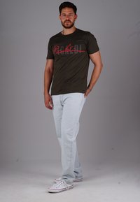 Mann steht mit einer Hand in der Tasche, trägt ein schwarzes Grafik-T-Shirt, hellblaue Jeans und weiße Sneakers vor einem schlichten Hintergrund.