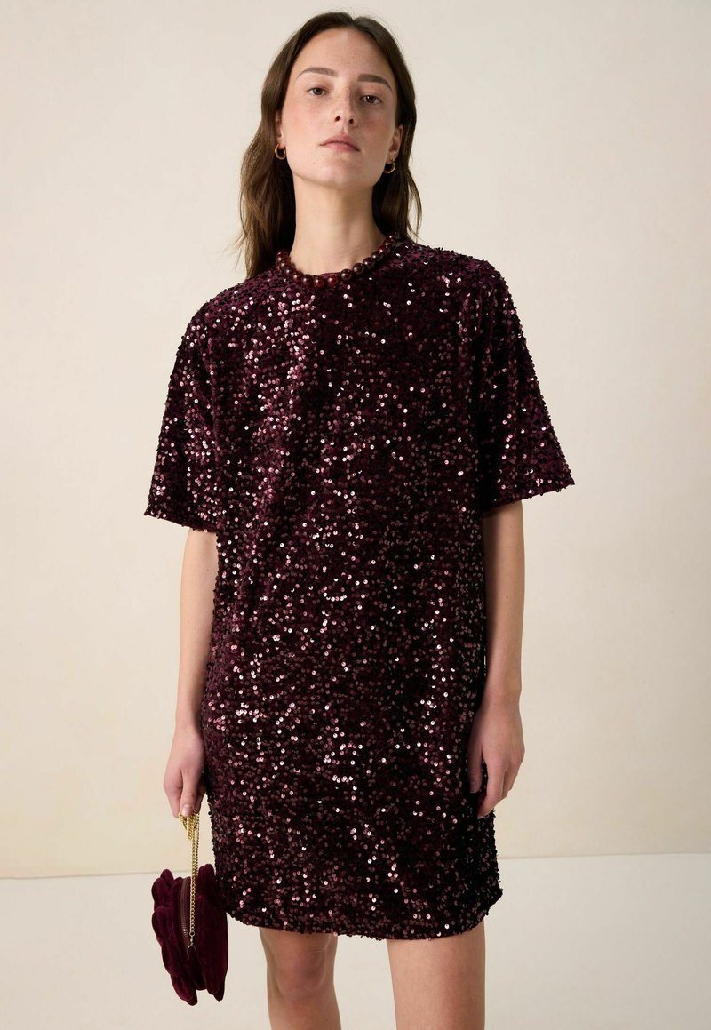 Mini-robe bordeaux à sequins avec manches courtes, encolure ronde et coupe ample. Le modèle tient un petit sac assorti.