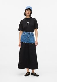 Fekete pólót, réteges maxi szoknyát farmer mini szoknyával, fekete cipőt és világoskék sapkát viselő nő, aki fehér háttér előtt áll.