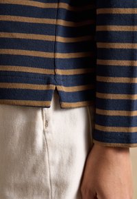 Chemise à manches longues rayée bleu marine et moutarde avec fente latérale portée sur un pantalon texturé couleur crème, montrant le bras et la main d'une personne.