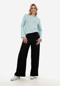 Donna sorridente con capelli biondi, che indossa una maglietta a maniche lunghe azzurro chiaro, pantaloni neri a gamba larga e sneakers bianche, in piedi con le mani sui fianchi.