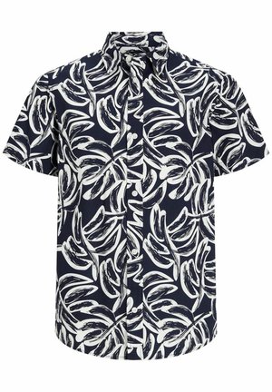 Garçon portant une chemise à manches courtes imprimée de feuilles en noir et blanc, un short gris retroussé, des chaussettes blanches et des baskets blanches avec des détails noirs.