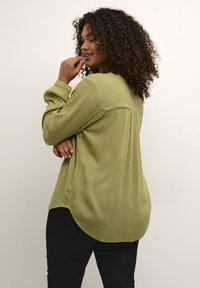 Camicia a maniche lunghe verde oliva con finitura texturizzata, orlo curvo e dettaglio del yoke posteriore. Indossata con pantaloni neri, mostrata di lato.