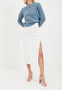 Hellblauer Strickpullover mit strukturiertem Muster, kombiniert mit einem weißen Midirock mit seitlichem Schlitz. Dazu werden hautfarbene Sandaletten mit hohem Absatz getragen.