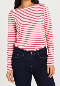 Camicia a righe rossa e bianca a maniche lunghe con scollo a barca, abbinata a jeans in denim blu scuro. La texture è morbida e aderente.