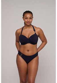 Vrouw met gevlochten haar draagt een donkerblauwe bikini met ringdetails, staand tegen een effen lichtgrijze achtergrond, met een zachte glimlach.