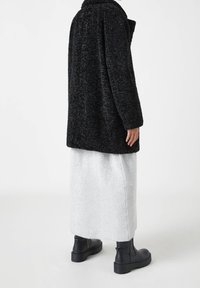 Manteau en fausse fourrure noire à la texture douce, coupe oversize et large col, associé à une jupe longue grise claire côtelée et des bottines noires.