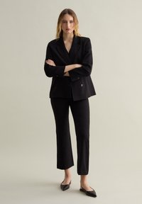 Blazer noir à double boutonnage avec revers larges, associé à un pantalon noir slim-fit et des mules noires à bout pointu. Tissu lisse et lignes épurées.