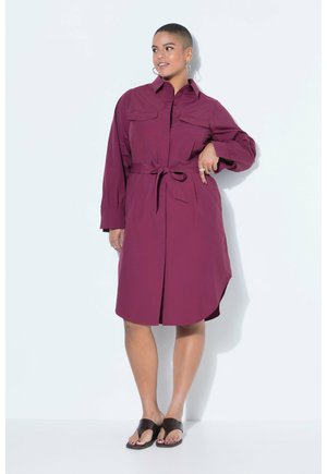 Kvinde med kort hår iført en lilla skjortekjole med lange ærmer og bælte samt sorte sandaler, stående foran en hvid baggrund.