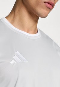 Λευκό αθλητικό μπλουζάκι με στρογγυλή λαιμόκοψη και υφασμάτινο κέντημα λογότυπου στο στήθος. Λείο ύφασμα με μινιμαλιστικό σχεδιασμό.