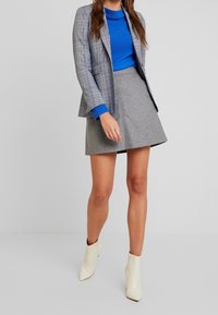 Grijze geruite blazer over een blauw ribbeltrui, gecombineerd met een grijze uitlopende rok en witte enkellaarzen, die een gestructureerd ontwerp en contrasterende kleuren toont.