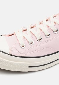 Sneaker di tela rosa chiaro con lacci bianchi, occhielli argentati e punta in gomma. Presenta una suola in gomma nera e dettagli testurizzati sulla parte superiore.