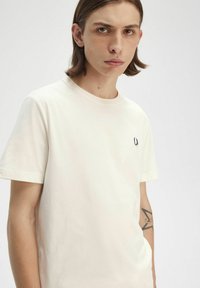 Camiseta de algodón color crema con cuello redondo y mangas cortas. Presenta un pequeño logo bordado en azul marino en el pecho. Textura suave.