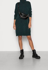 Vestito a collo alto in maglia verde, lunghezza fino a metà coscia, abbinato a una borsa a tracolla con catena nera e stivali neri con suola spessa.