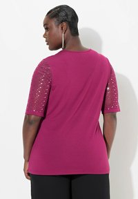Fuchsia t-shirt med korte, gennemsigtige ærmer prydet med sølvfarvede palietter. Har en rund halsudskæring og en afslappet pasform. Glat stofstruktur.