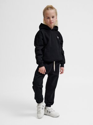 Blond dziecko stoi, ubrane w czarną bluzę z kapturem i pasujące spodnie dresowe, białe trampki, na jednolitym jasnym tle.